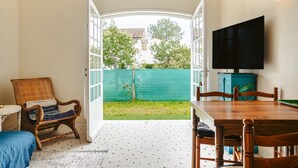 An open-plan living area with a dining table, a television mounted on the wall, a wicker chair, and a view of the backyard.
