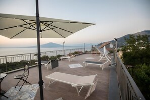 Une terrasse extérieure avec vue sur la mer, un grand parasol et des chaises longues.