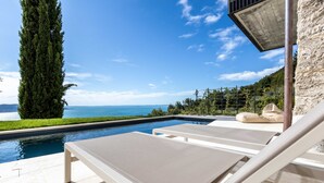 Ein Poolbereich mit Liegestühlen, einem hohen Baum und Blick auf das Meer.