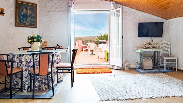 An open-plan living area with a dining table, chairs, a television, and a view through the open door.