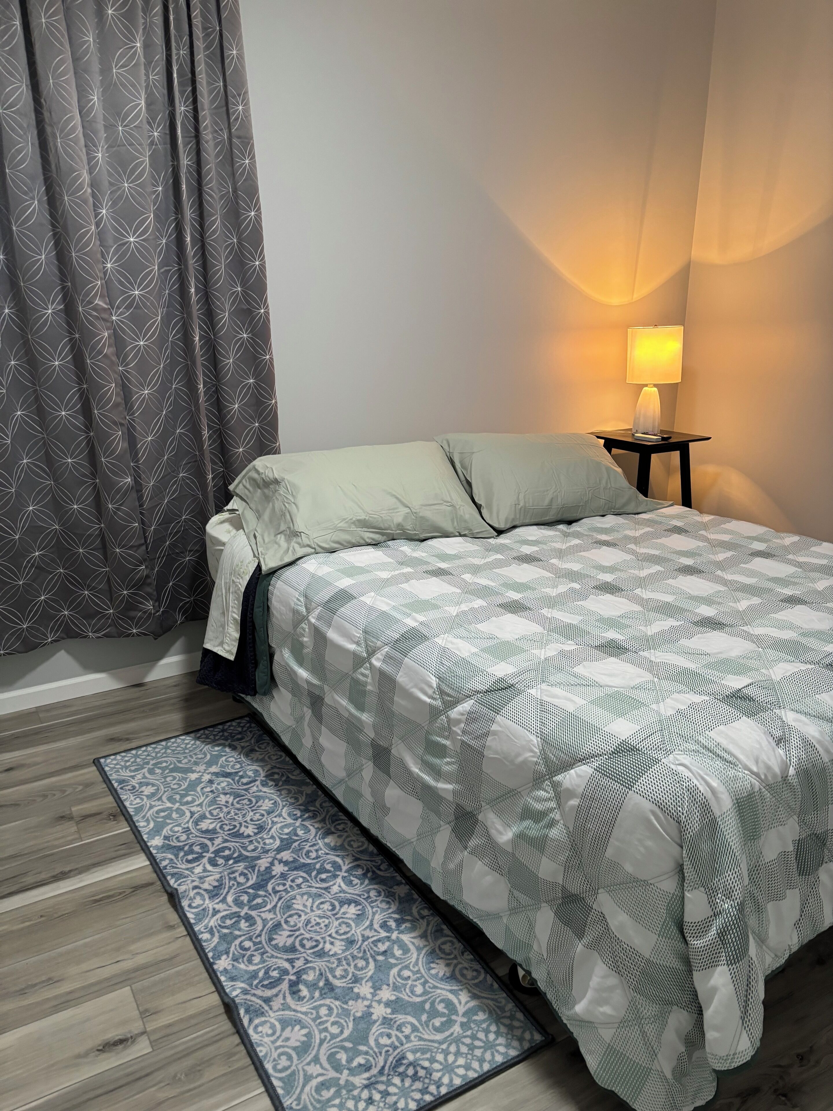 Een slaapkamer met een bed, een nachtkastje met een lamp, een kamerkleed met een patroon en een raam met gordijnen.
