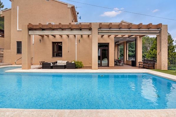 Una piscina con un porche techado, mobiliario exterior y un edificio de fachada color crema.