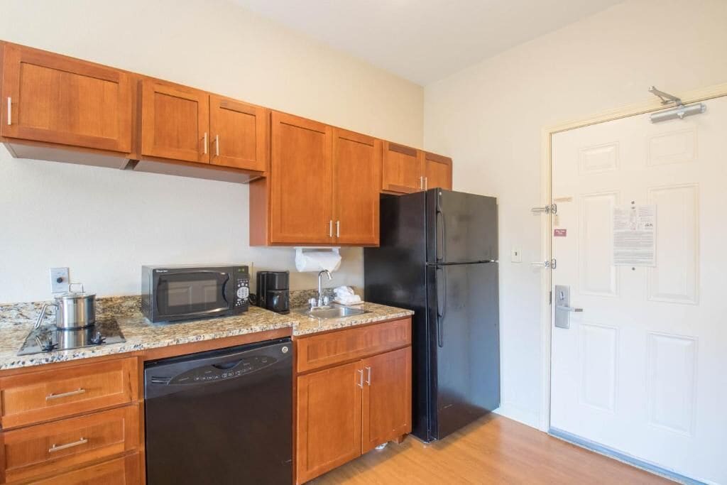Cocina compacta con armarios de madera, un refrigerador negro, microondas, cafetera y lavavajillas.