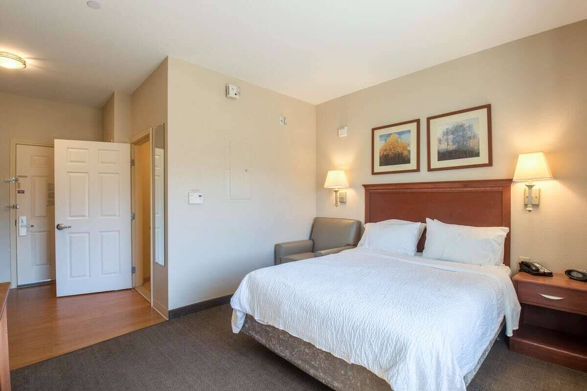 A hotel room with a large bed, two bedside lamps, a nightstand, a chair, and a door leading to another room.