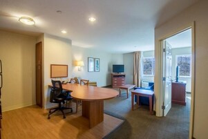 Quarto de hotel com mesa de jantar de madeira, cadeiras, televisão, escrivaninha e janela com cortinas.
