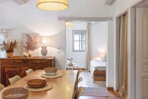 A dining area with a wooden table, chairs, a lamp, and a view of a bedroom with a bed and curtains.