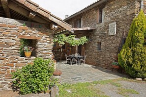 Edificio de piedra con un patio que incluye una mesa, sillas, una planta en maceta y un muro de piedra.