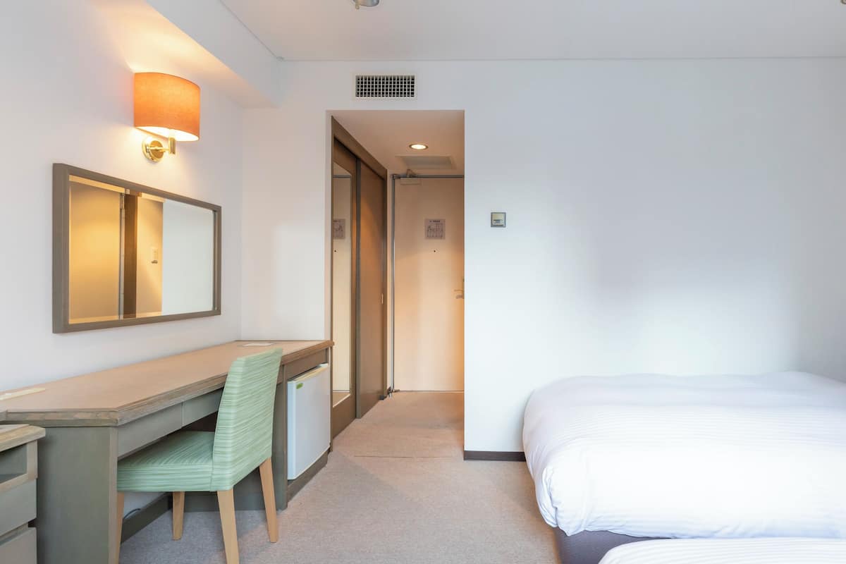 A hotel room with a desk, chair, bed, and a wall-mounted lamp.