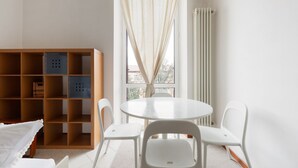 A room with a round table, white chairs, a wooden bookshelf, and a window with curtains. - GuestHost - Gambara M1 White Functional Appt x4 (Milano)