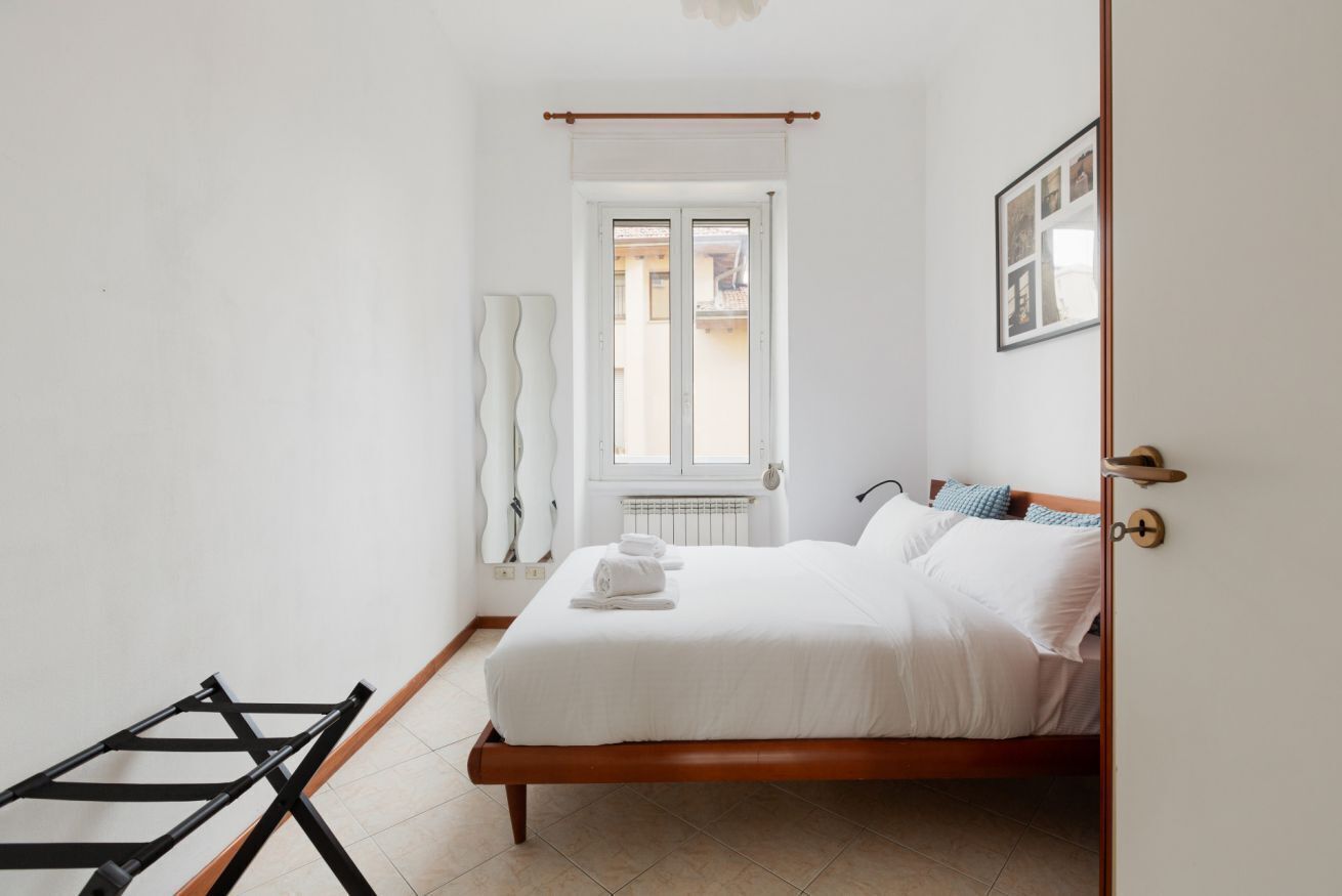 A bedroom with a wooden bed, white bedding, a radiator, a window with curtains, and a door with a handle.