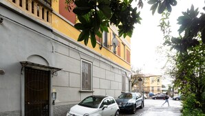 A narrow street with parked cars and a building with a large door and a satellite dish. - GuestHost - Gambara M1 White Functional Appt x4 (Milano)