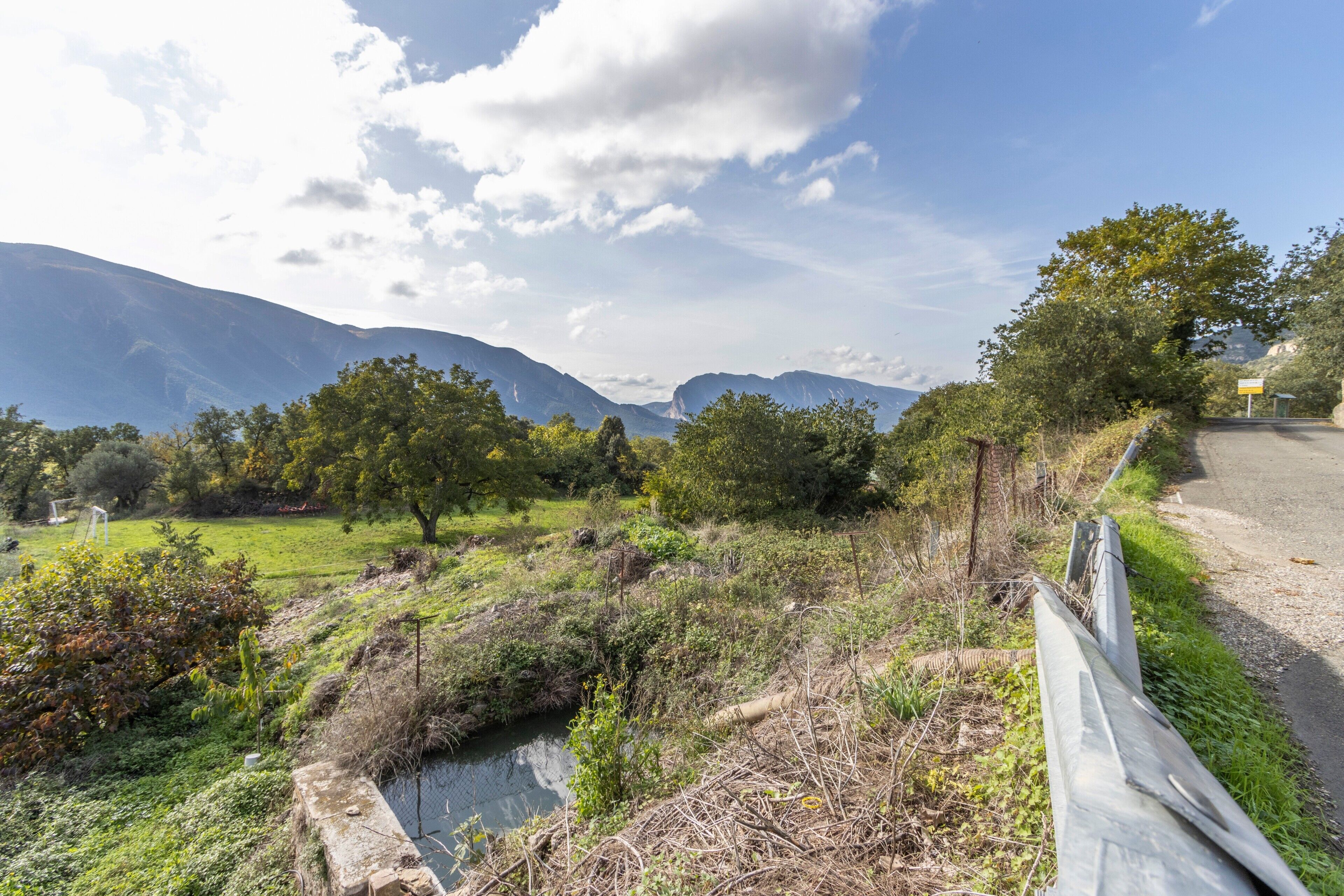 Un paisaje rural con una carretera, un arroyo y montañas al fondo.