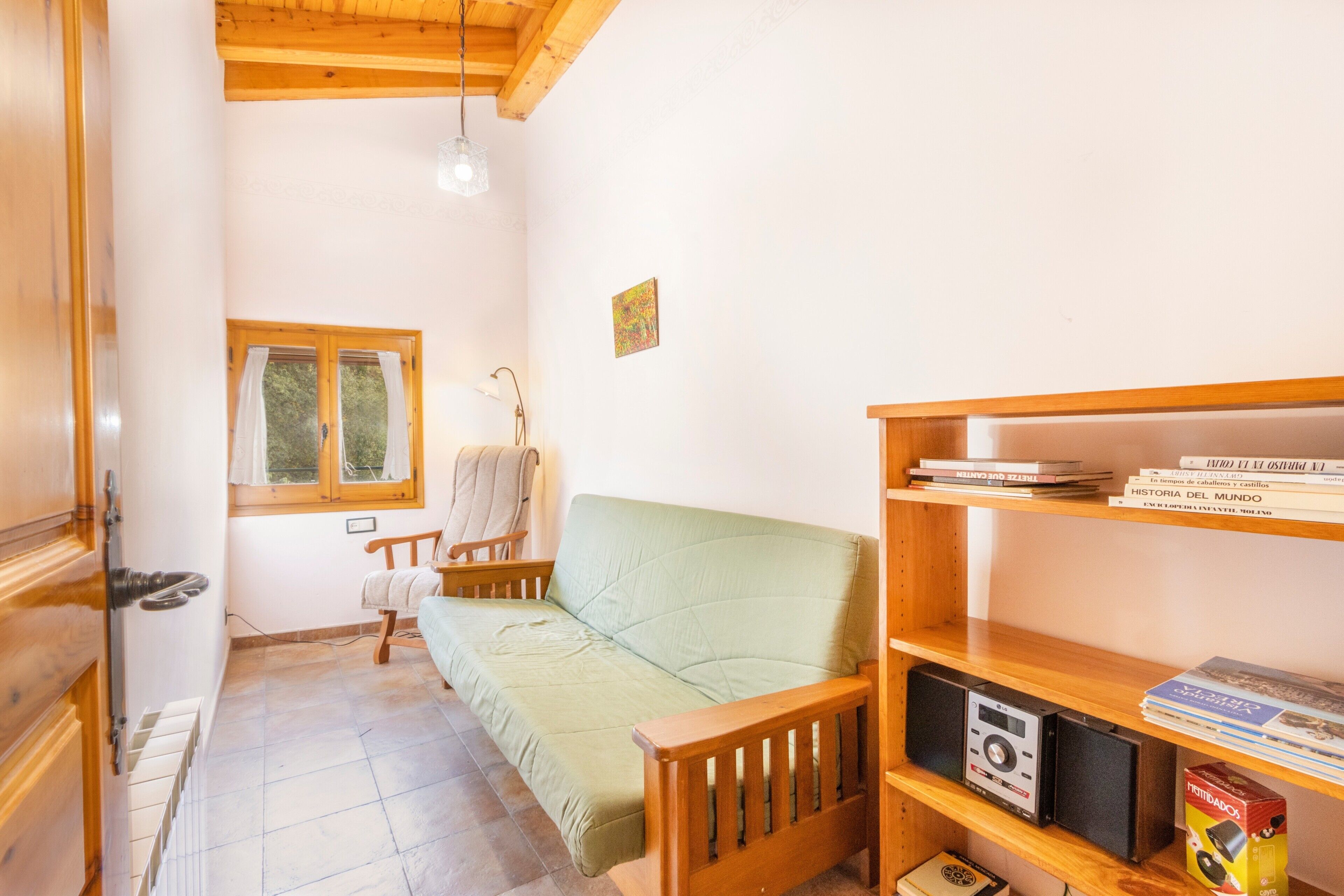 Una habitación acogedora con un banco de madera, un sofá verde, una mesita con una lámpara, una ventana con cortinas y una estantería con libros y una cámara.