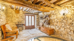 Una habitación rústica con paredes de piedra, un banco de madera, una cesta de mimbre, un cofre de madera y una mesa de cristal.