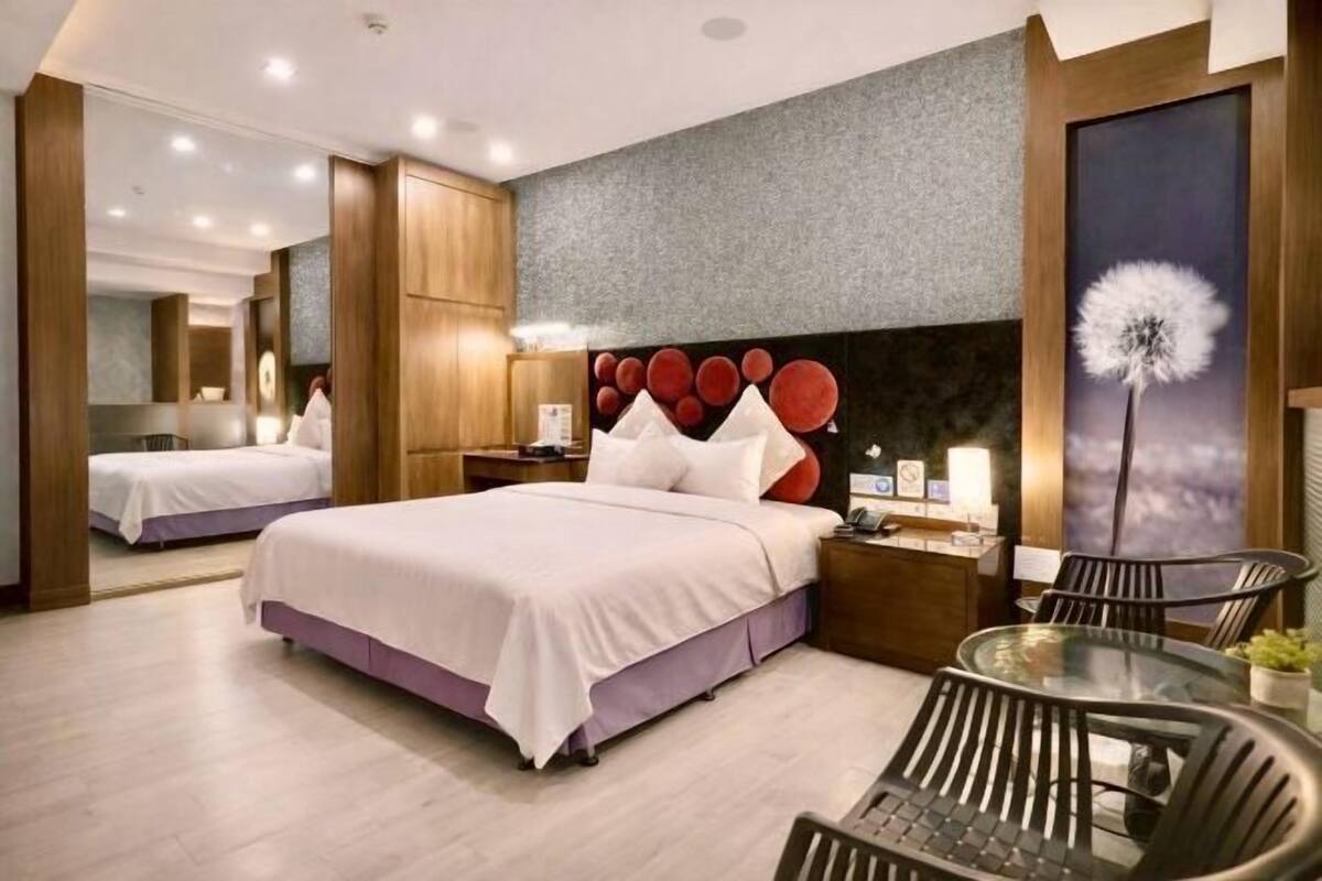 A modern hotel room with a large bed, a bedside table with a lamp, a small round table with chairs, and a large mirror.