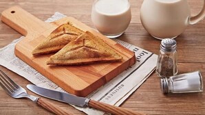 A wooden cutting board with two triangular sandwiches, a glass of milk, a milk jug, a salt shaker, a knife, and a fork on a wooden table with a newspaper underneath. - Wei Fengqing Motel (Tainan) (Tainan)