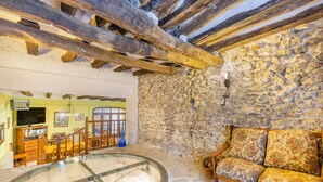 A rustic interior with a stone wall, wooden beams, a glass table, and a floral sofa.