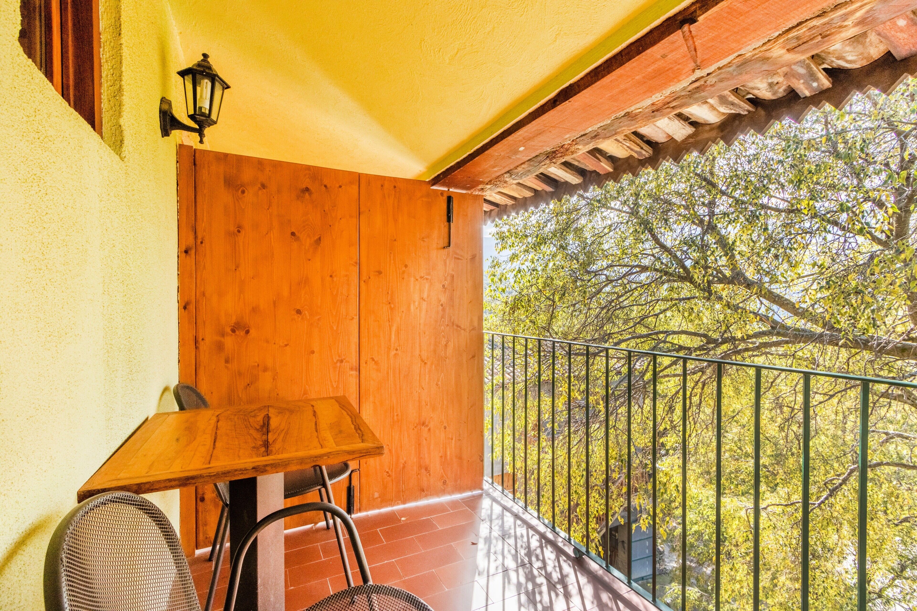 Een balkon met een houten tafel en stoelen, een metalen hekwerk, en uitzicht op bomen.