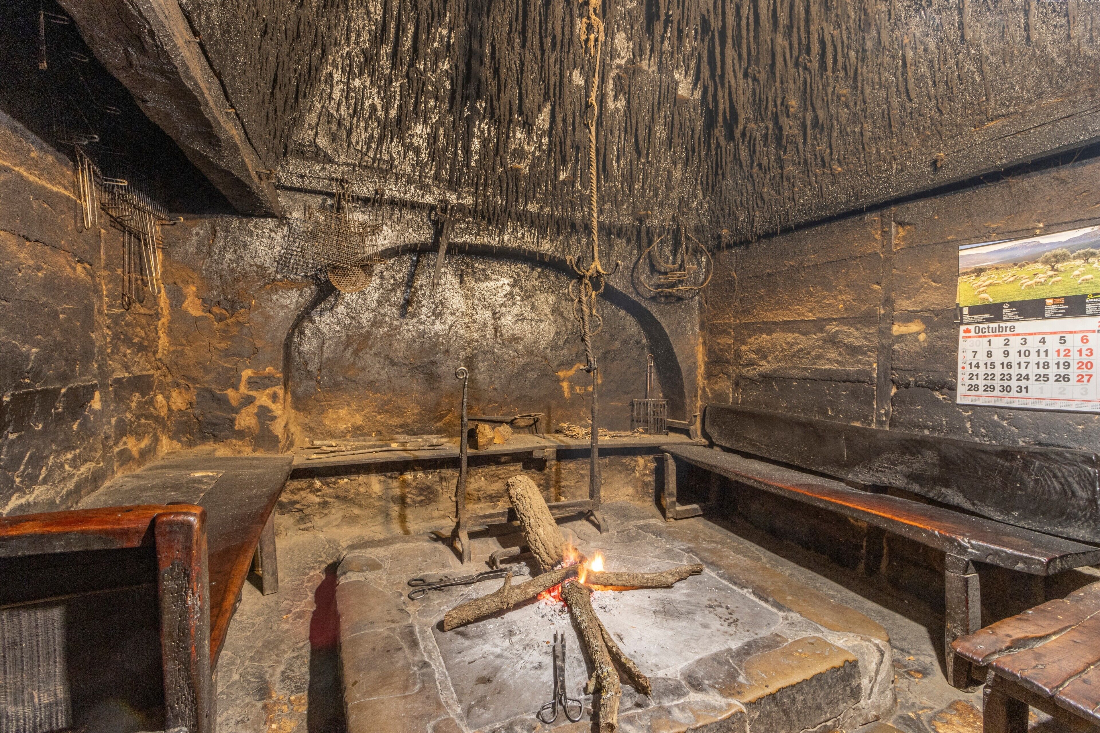 Un interior de piedra con una chimenea, bancos de madera y un calendario en la pared.