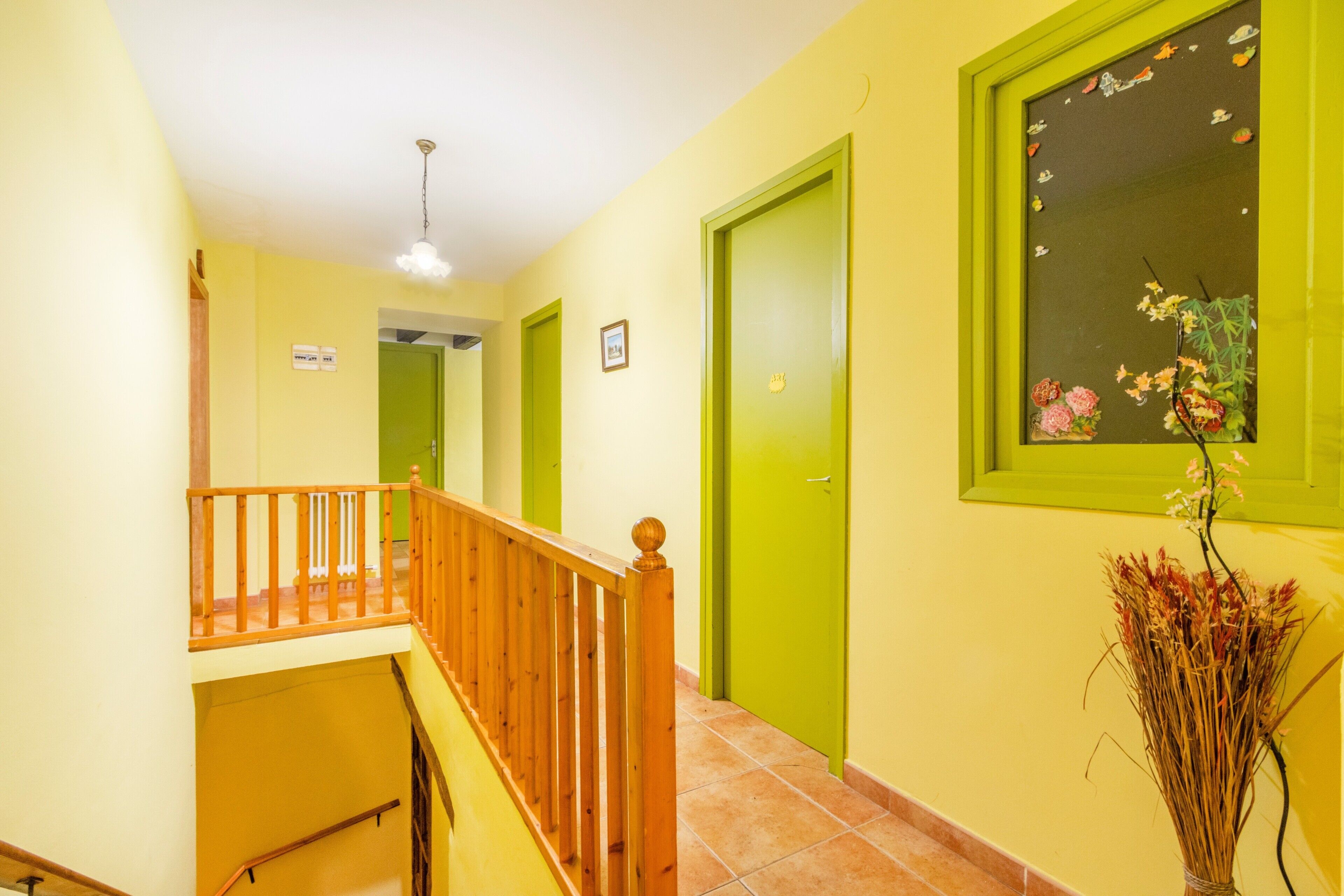 A hallway with a wooden railing, green doors, and a vase with dried plants.