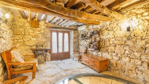 A rustic stone-walled room with a wooden bench, a wicker basket, a wooden chest, and a glass table.