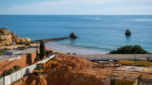 Un chantier de construction situé sur la côte, avec une plage, des affleurements rocheux et une excavatrice jaune.