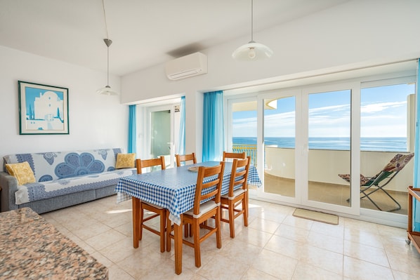 En matplats med ett bord för fyra personer, en soffa och utsikt över havet genom en skjutdörr i glas.
