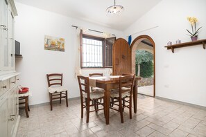 A dining area with a wooden table and chairs, a window with curtains, a painting on the wall, and a wooden door leading outside.