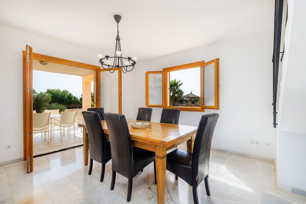 Un comedor con mesa de madera, sillas negras, una araña y vistas a través de un ventanal abierto.