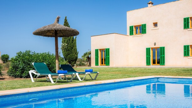 Una piscina con sillas de descanso y una sombrilla de paja, un edificio con contraventanas verdes y un cielo azul despejado.