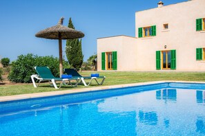 Una piscina con sillas de descanso y una sombrilla de paja, un edificio con contraventanas verdes y un cielo azul despejado.
