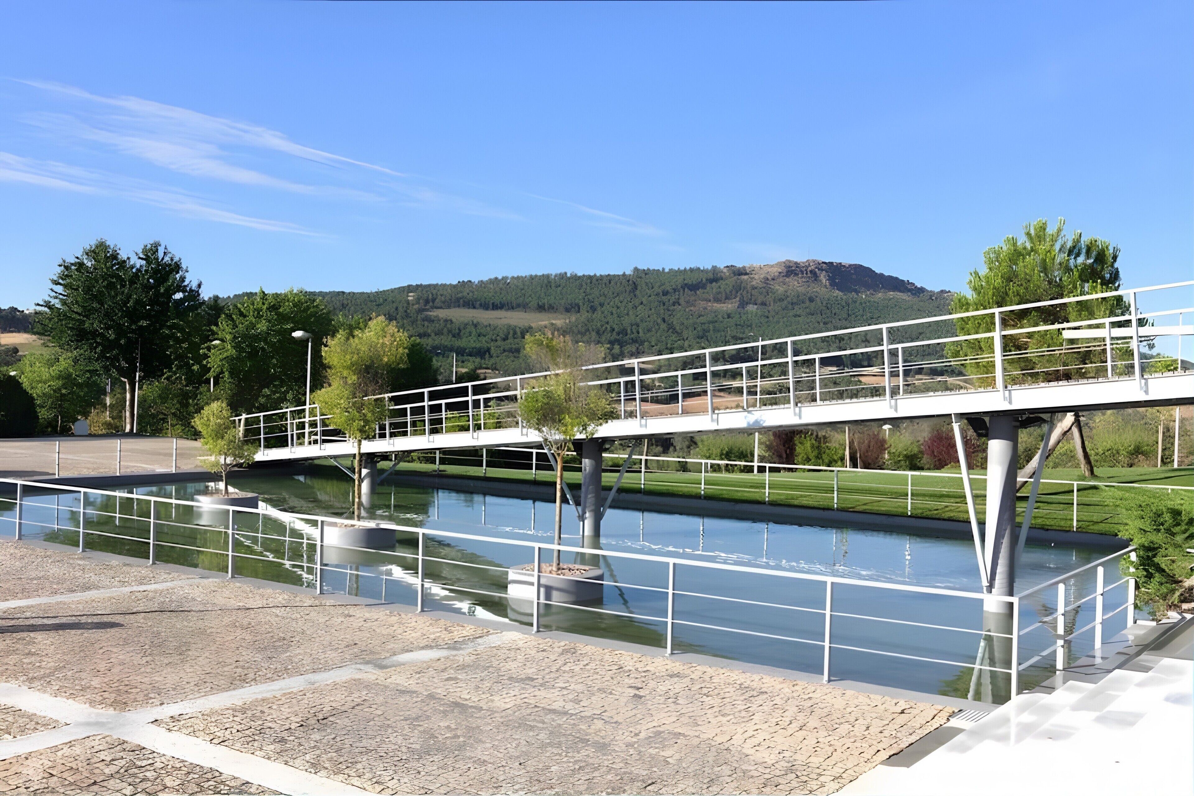 A bridge over a canal with a clear sky, trees, and a hilly landscape in the background.