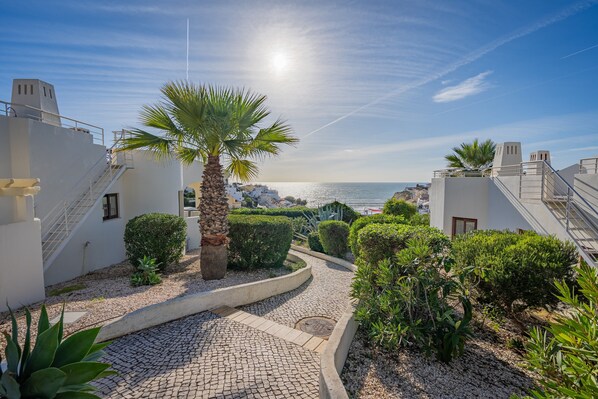 A coastal building with a paved pathway, palm trees, and a view of the sea. - Apartment 'Norah’s House' with Shared Pool, Wi-Fi and Air Conditioning (Carvoeiro)