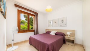A bedroom with a bed, a bedside table with a lamp, a standing fan, and a window with curtains.