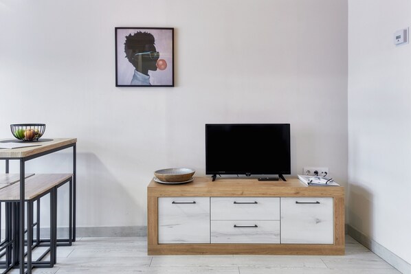 A modern living room with a TV on a wooden cabinet, a framed picture on the wall, and a small table with a bowl of fruit.