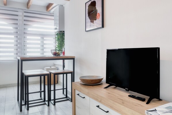 A modern kitchen with a wooden table, two stools, a large black TV, and a bowl on the counter.