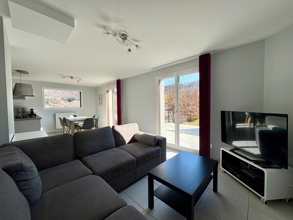 A modern living room with a sofa, a coffee table, a TV on a stand, a dining area with a table and chairs, and a large window with a view of trees.
