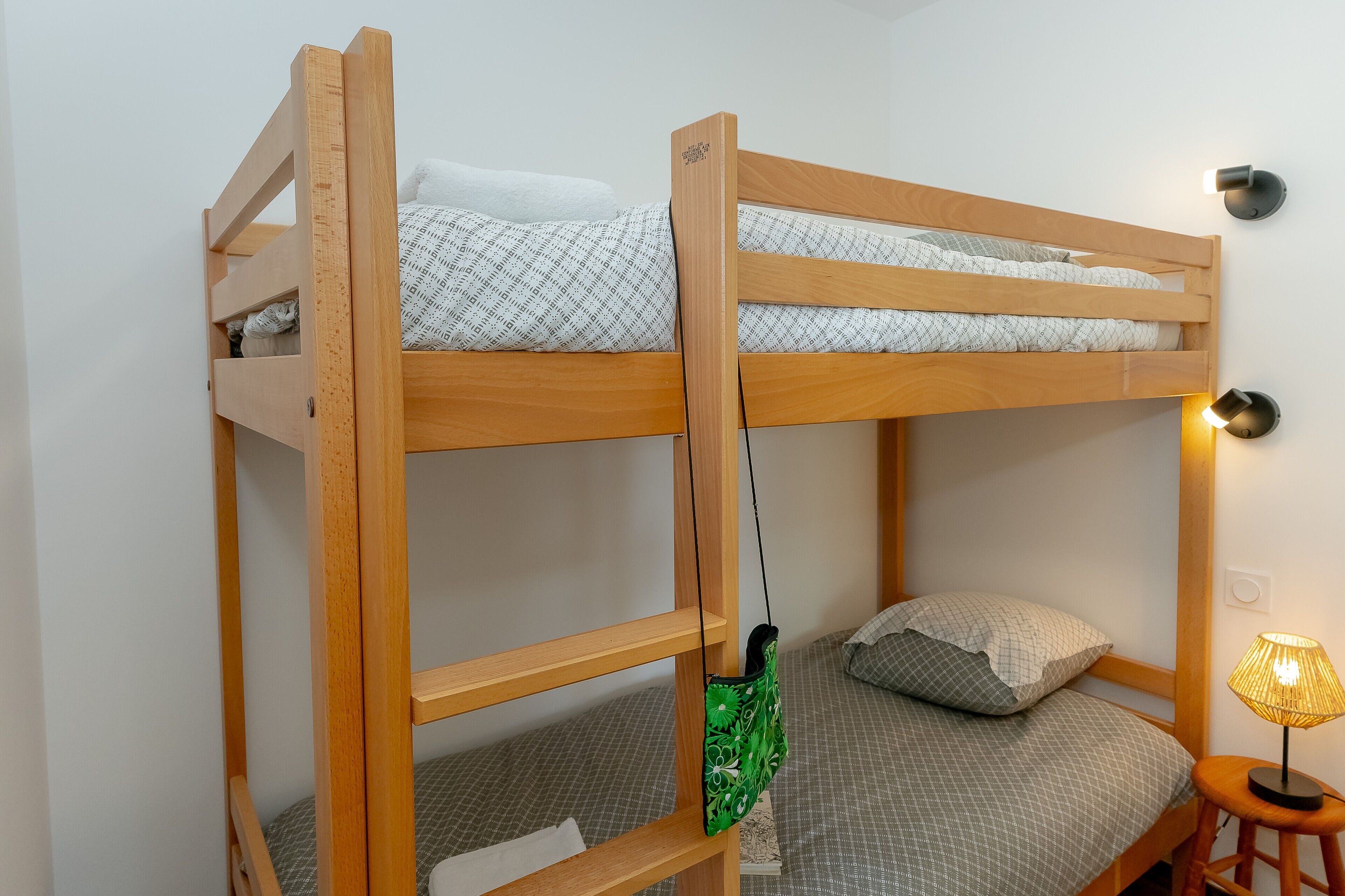 Ein Stockbett mit einer grünen Tasche an der Seite, ein kleiner Hocker und eine Wandlampe.