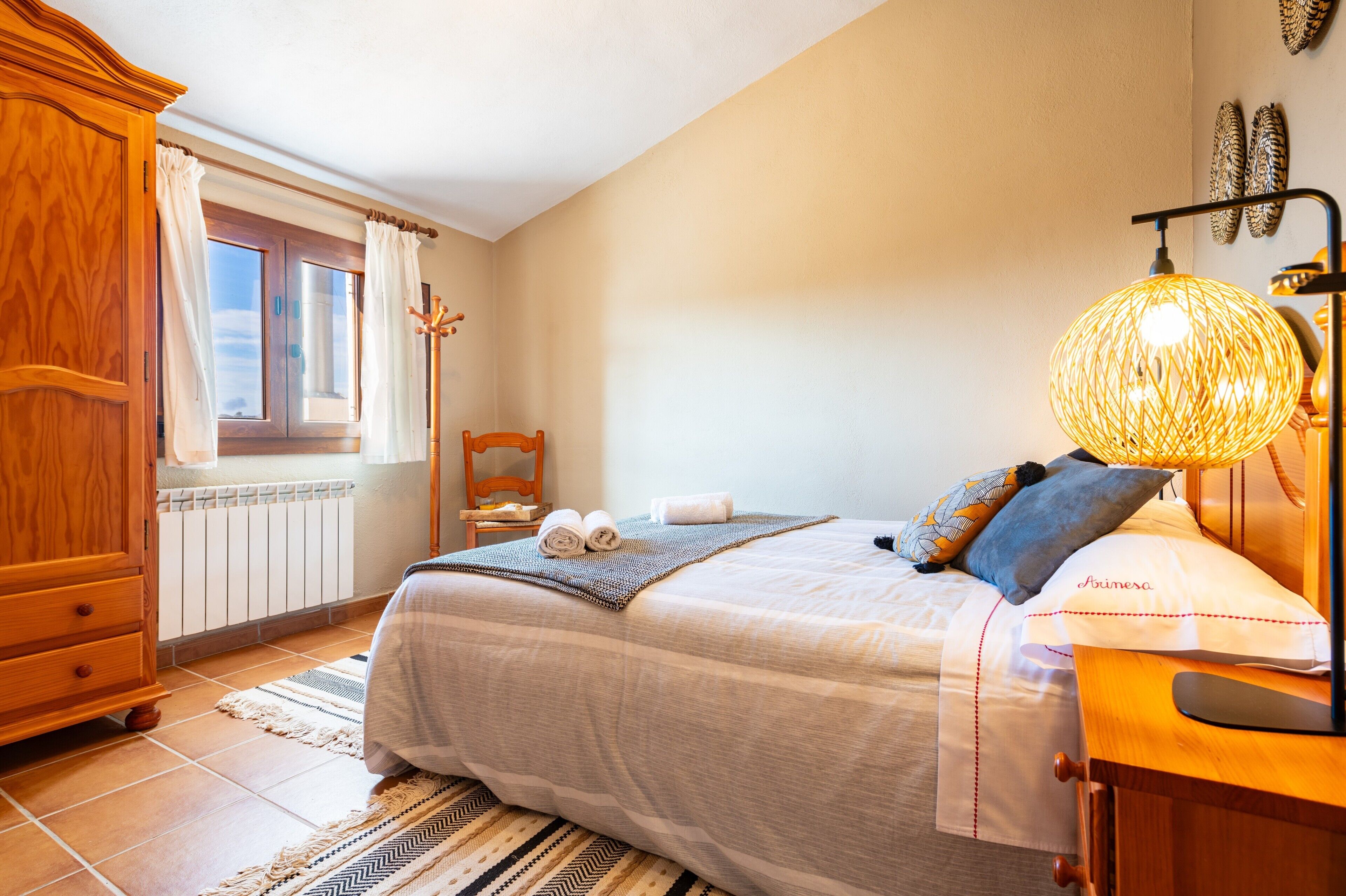 Et soveværelse med en seng, et træskab, et vindue med gardiner, et natbord med en lampe og en hylde monteret på væggen.