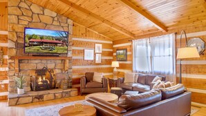 Ein Blockhaus-Interieur mit Kamin aus Stein, Ledersofa, Holzwänden und einem an der Wand befestigten Fernseher.