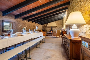 Uma zona de refeições rústica com uma mesa comprida posta para uma refeição, paredes de pedra e vigas de madeira aparente.