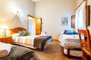 A hotel room with two beds, a wooden headboard, a bedside table with a lamp, a chair, and a door leading to another room.