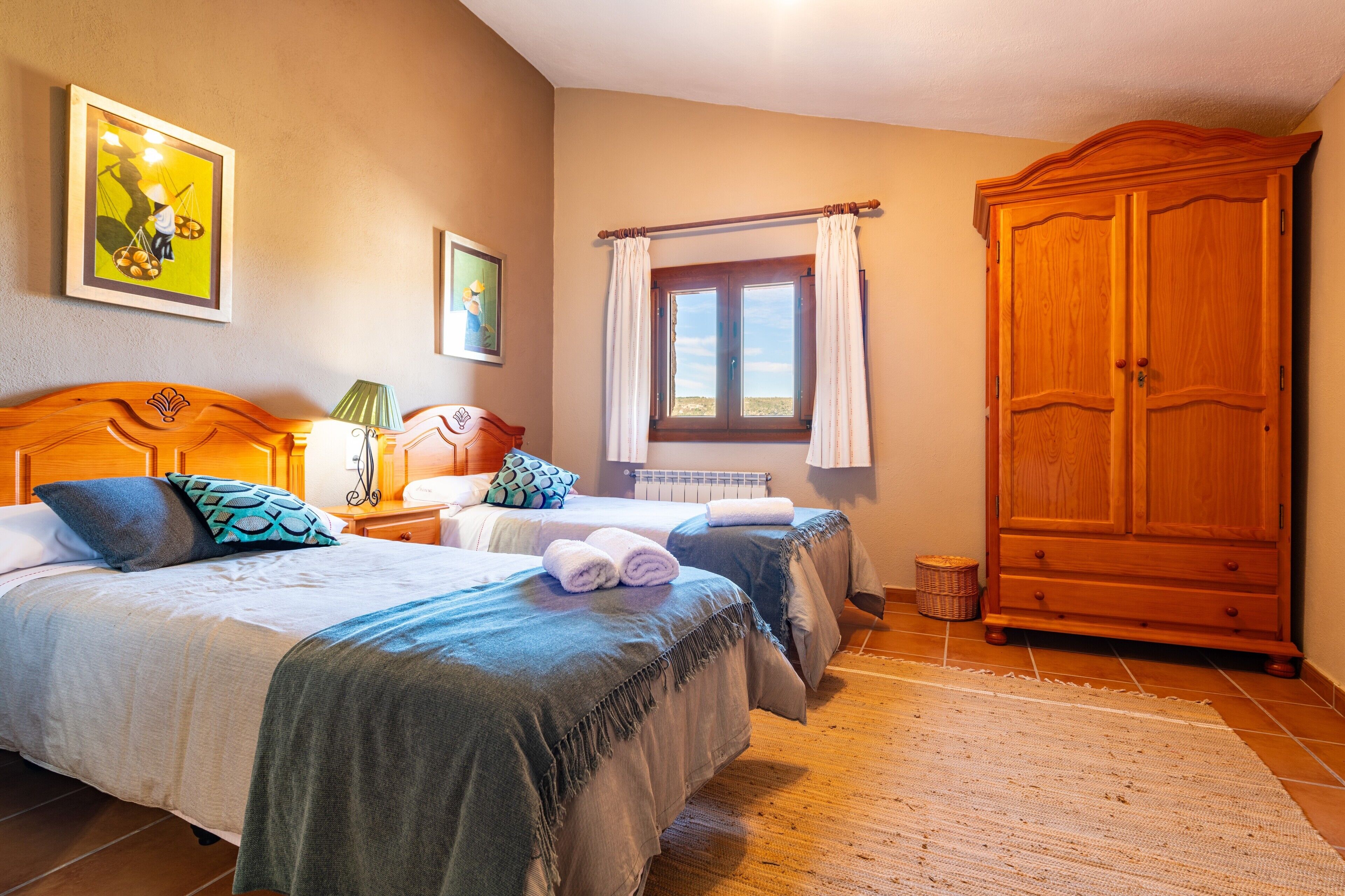 A hotel room with two beds, a wooden wardrobe, a window with curtains, and artwork on the walls.