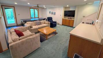 A living room with a sofa set, a coffee table, a ping pong table, a TV, and a kitchen area.
