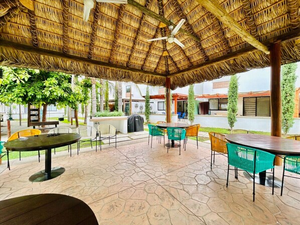 An outdoor patio area with round tables and chairs, a thatched roof, and a stone floor.