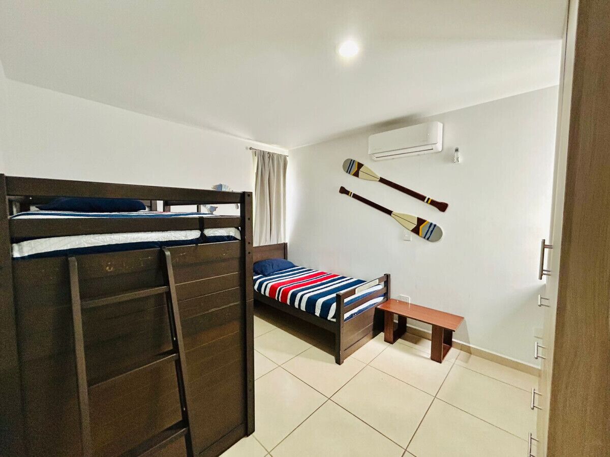 A bedroom with a bunk bed, a single bed with a striped blanket, a wooden bench, and two paddles on the wall.