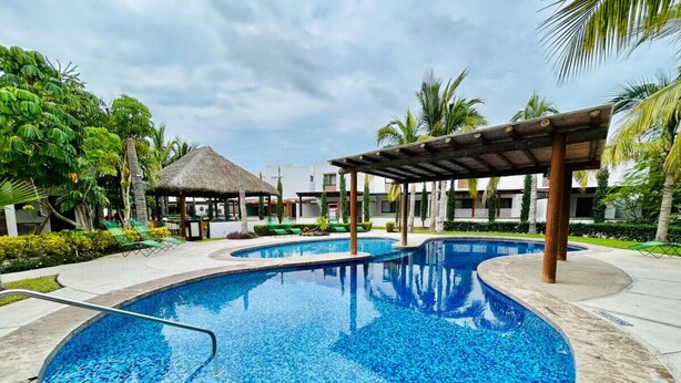 A large outdoor swimming pool with a wooden deck and lounge chairs, surrounded by palm trees and a thatched-roof structure. - Casa en privada frente a la alberca by PartnerHost (Mazatlán)