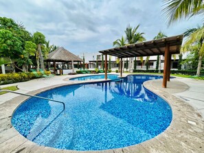 A large outdoor swimming pool with a wooden deck and lounge chairs, surrounded by palm trees and a thatched-roof structure.