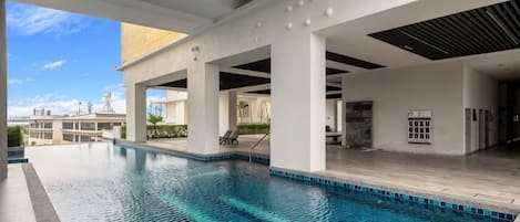 A swimming pool with clear blue water, surrounded by a covered area with white pillars and a tiled floor.