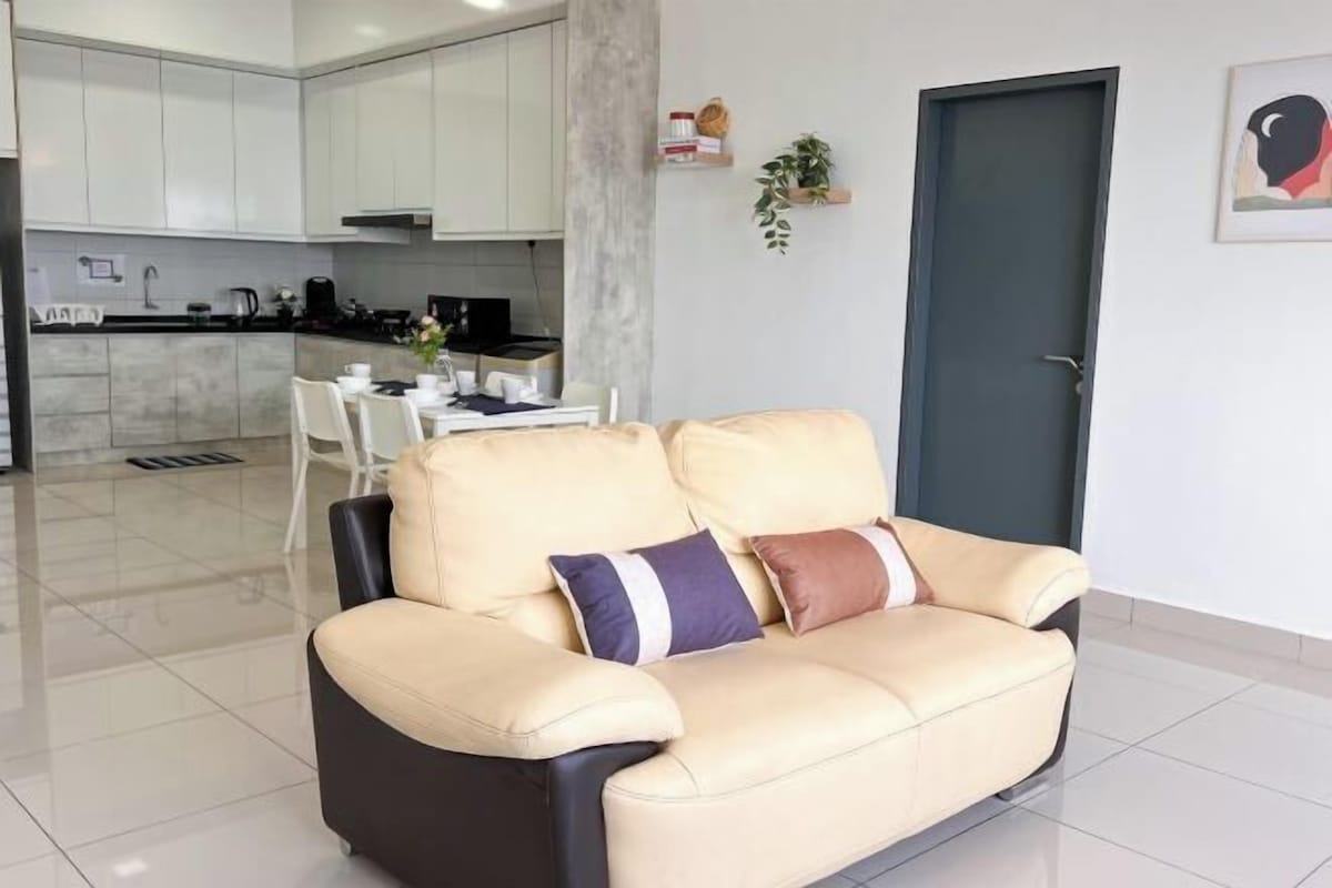 A modern living room with a beige sofa, a dining table with chairs, a kitchen in the background, and a dark door.